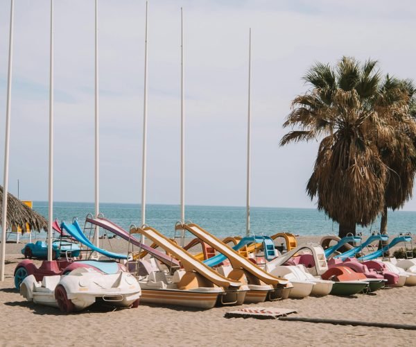 pexels-photo-16753341-16753341 Vibrant pedal boats on a sunny Málaga beach with palm trees and clear skies.